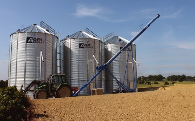 un tracteur avec vis sur chariot devant 3 silos à grain de stockage